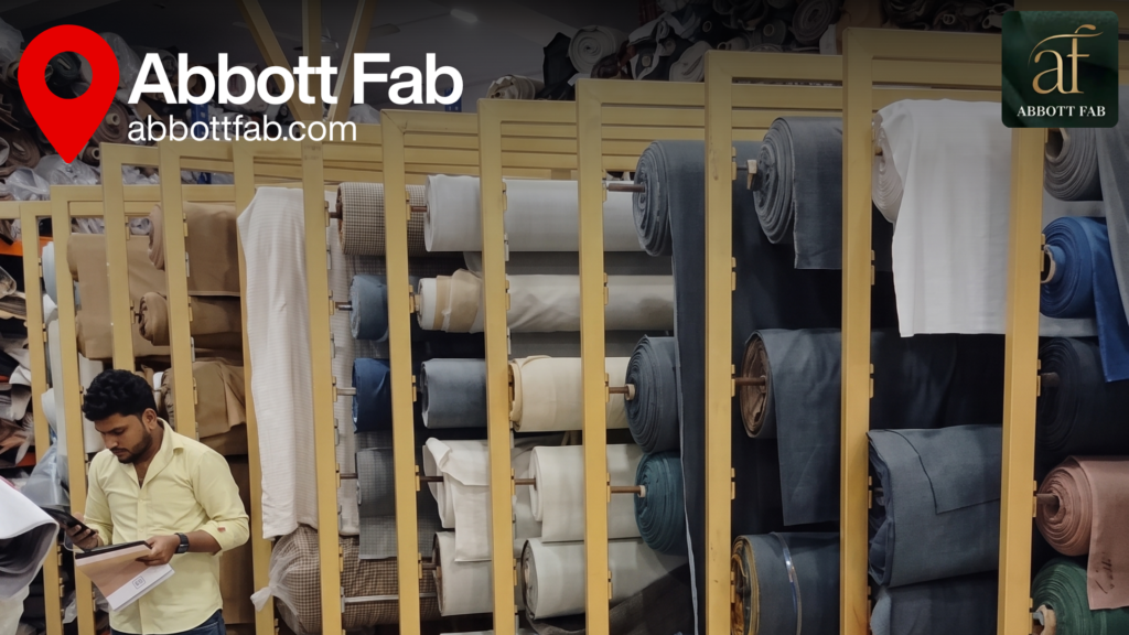 ABBOTT FAB showroom and warehouse image featuring a red location pin, the Abbott Fab brand logo, website url abbottfab.com, and a man holding sample folders while inspecting rows of yellow metal storage racks filled with rolled upholstery fabrics and bolts in neutral, blue, and earth tones