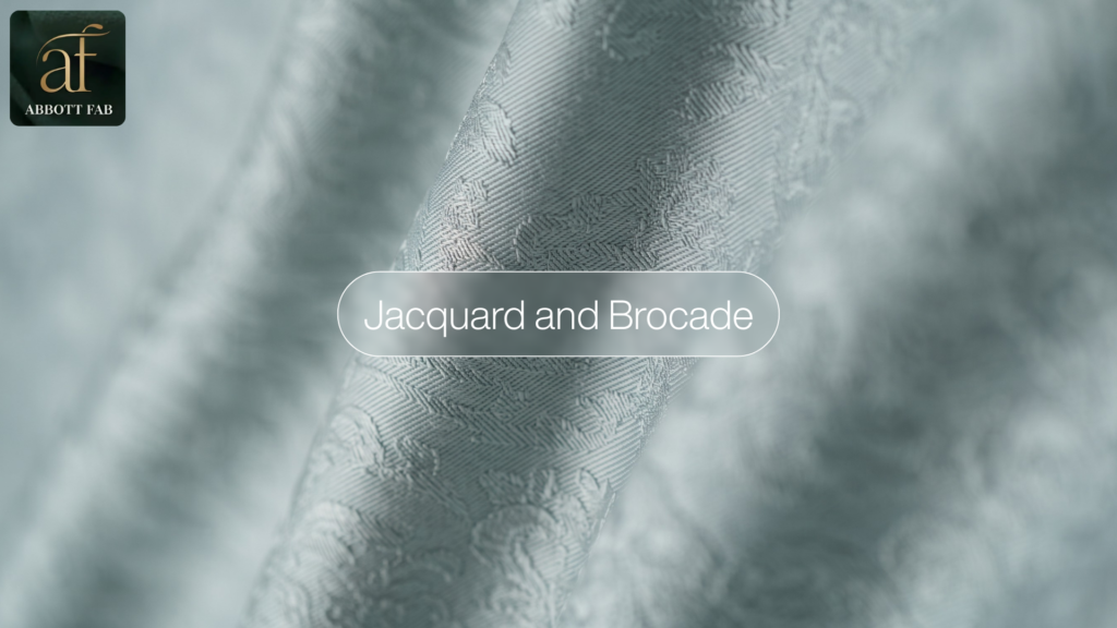 ABBOTT FAB product showcase image featuring the Abbott Fab brand logo, a centered badge reading Jacquard and Brocade, and a close-up photograph of light grey patterned fabric with intricate woven motifs in soft folds.